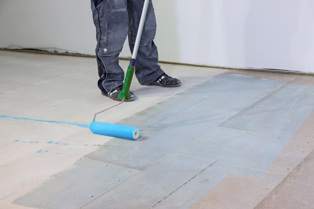 UZIN_Application Image_PE360 Plus_Renoplan System_2020-10 Man ser en arbejders underben og fødder, mens han med en lang malerulle påfører en lyseblå primer eller maling på et trægulvs underlag. Personen har mørke arbejdsbukser og sorte sneakers; rullen efterlader et vådt blåt lag på gulvbrædderne i et rum med hvide vægge. Billedet fokuserer på gulvforberedelsen, værktøj og malingssprøjt.