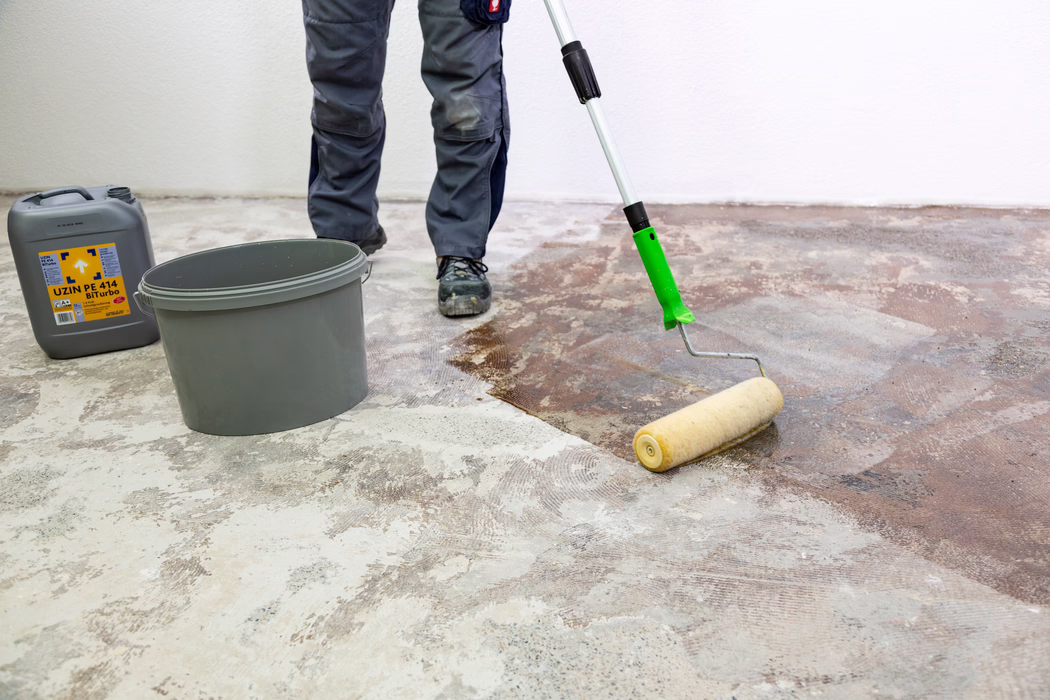 UZIN_Application Image_PE 414_9406_2018-11 En arbejders underben og fødder kan ses, mens han bruger en langskaftet malerulle til at påføre en brun primer eller lim på et bart betongulv. En grå plastikspand og en grå kanister mærket "UZIN PE 414 Biturbo" står i nærheden ved en hvid væg. Arbejderen bærer grå arbejdstøj og sorte sikkerhedssko; gulvet viser pletter af gammel lim og slibet beton, mens rullen fordeler væsken jævnt for at forberede underlaget.