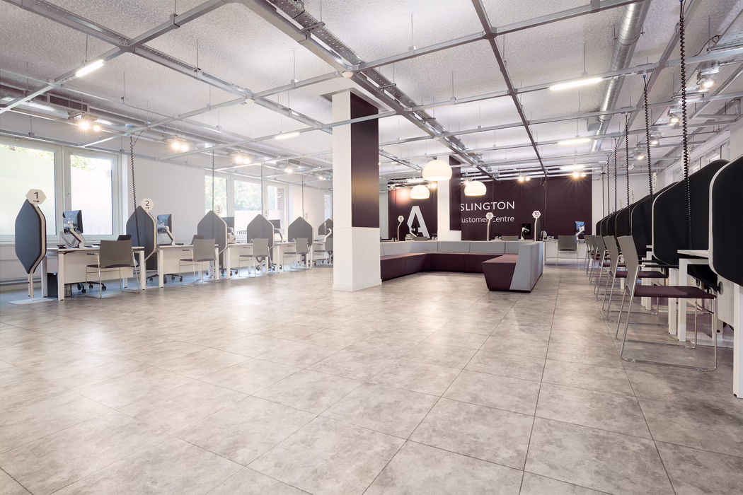 A bright, modern customer service hall with a wide open floor and light grey tiled flooring. Along the left and right walls are rows of white service desks with high-backed dark privacy panels and numbered signs above each workstation; each desk has a computer monitor and a simple visitor chair. The center of the room features a large square column and a modular purple-and-grey seating area with pendant lamps hanging above. The ceiling shows an exposed structural grid with suspended lighting rails and visible ventilation ducts, giving a contemporary industrial look. Large windows along the left wall admit natural daylight; the space is clean, minimal and currently unoccupied.
