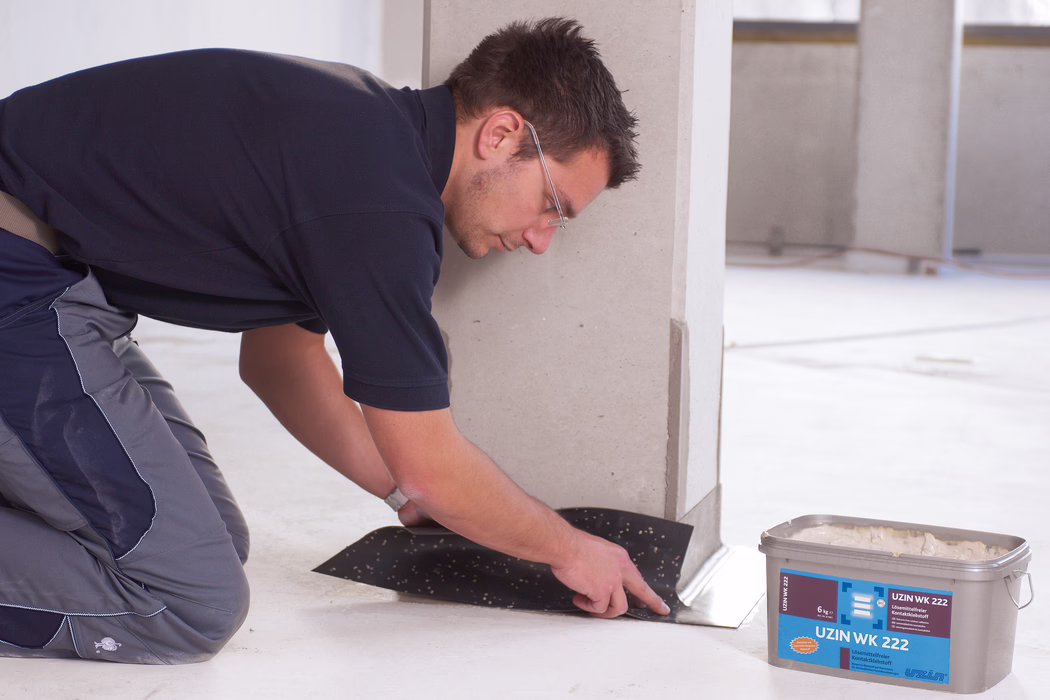 UZIN_Application_Image_WK 222_VA_3_2018-03 A construction worker wearing safety glasses and work clothes kneels inside a clean, unfinished interior and presses a black adhesive membrane or underlayment at the base of a light-colored wall or column. He smooths the material with his hands along the floor edge. Nearby on the floor is a plastic container labeled 'UZIN WK 222' containing adhesive. The scene shows a concrete or screed floor with minimal background details.