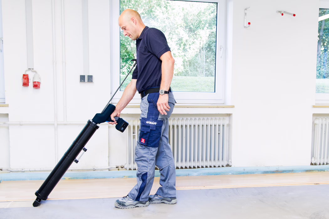 En servicemedarbejder bruger et langt sort gulvrengøringsværktøj indendørs, står foran et vindue og en radiator, iført en mørkeblå polo, grå arbejdstøj og sikkerhedssko.