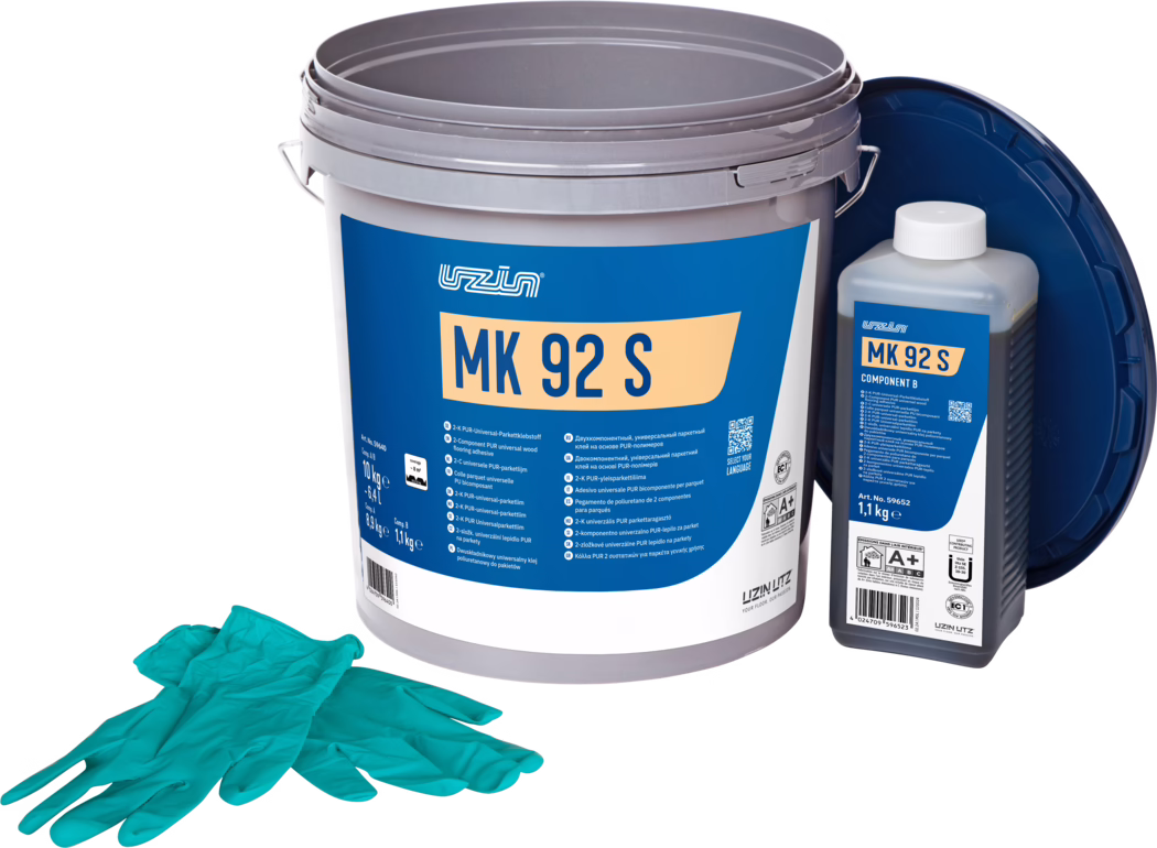 UZIN_Dummy_MK 92 S-10kg_2024-09 Studiefotografi af et parketlimsæt: en stor grå plastspand med en markant blå etiket med 'MK 92 S', til højre en mindre rektangulær flaske mærket 'MK 92 S Component B' med hvid hætte, og et mørkeblåt låg lænet bagved. Foran ligger et par turkise engangshandsker. Genstandene er arrangeret på en hvid baggrund med jævn belysning, hvilket fremhæver emballagedetaljer og produktoplysninger til gulvinstallation.
