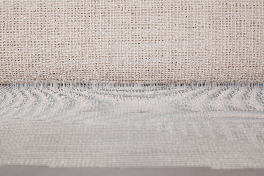 Nærbillede af et neutralt beige vævet stof med en åben, gitterlignende vævning. Øverst ses en rullet kant, der skaber en fin skyggekant; ujævne tråde og variationer i naturlige fibre giver et taktilt, rustikt udseende. Blødt, ensartet lys fremhæver teksturen og den subtile dybde.