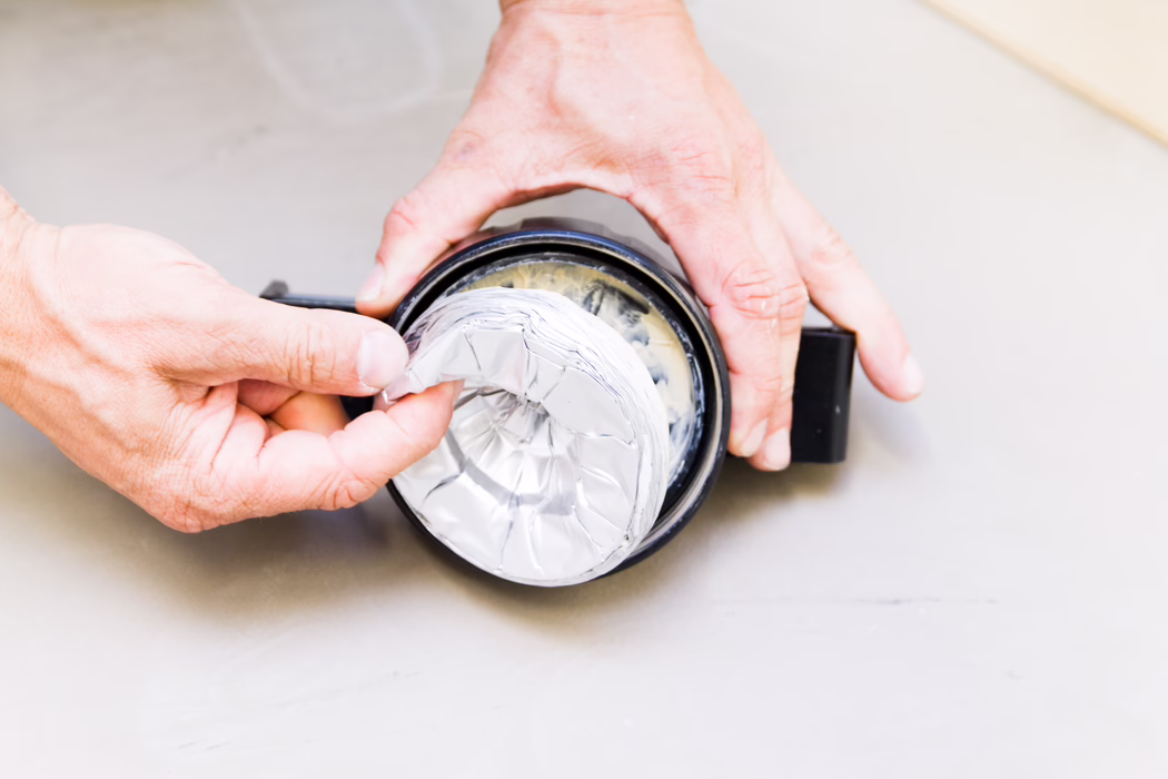 UZIN_Application_Image_ATD 100_32_2012-08 Close-up of two adult hands holding and assembling a small round black container while smoothing or removing a crumpled sheet of aluminum foil placed inside. The image is taken from above on a pale gray surface; the foil reflects light with visible creases and a ring of adhesive or sealing compound lines the interior rim. The hands show natural skin texture and light wrinkles, suggesting a DIY repair or sealing task with neutral, well-lit colors and a shallow depth of field.