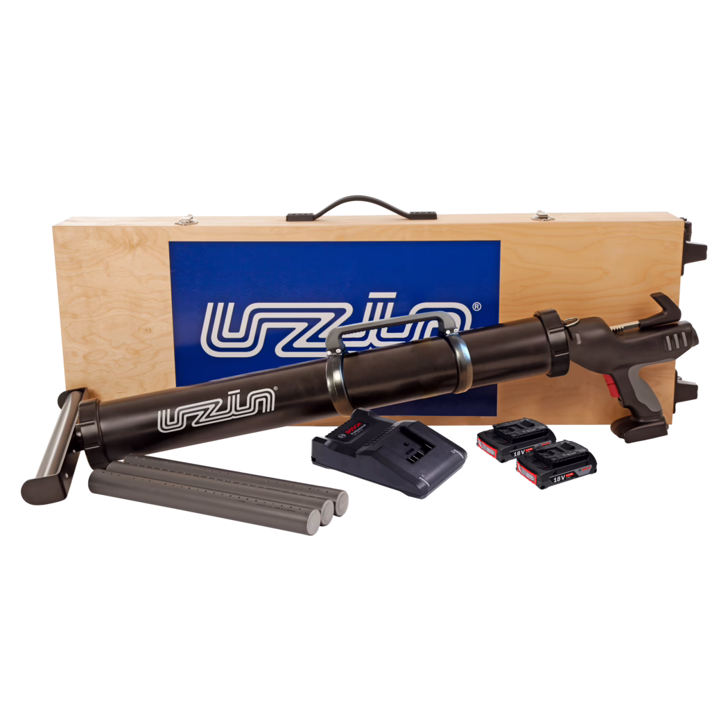 UZIN_Dummy_ATD 100 NEW_2021-04 Wooden transport case with a black cordless caulking/adhesive gun and accessories: the top of a light plywood case with a black handle and a blue logo panel is visible behind a long black battery-powered caulking gun. In front of the case are three grey cartridge tubes, a battery charger base and three 18V lithium-ion battery packs. The gun features a pistol grip with grey and red accents, metal strap clamps around the cartridge barrel and a trigger assembly. Clean studio composition on a white background.